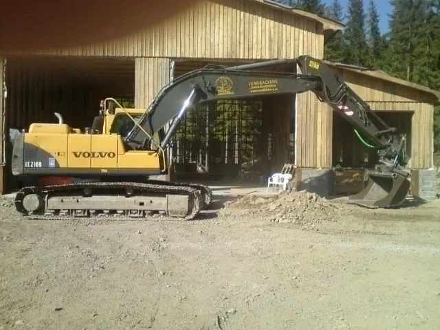 Gul grävmaskin på en grusplan vid skogskanten