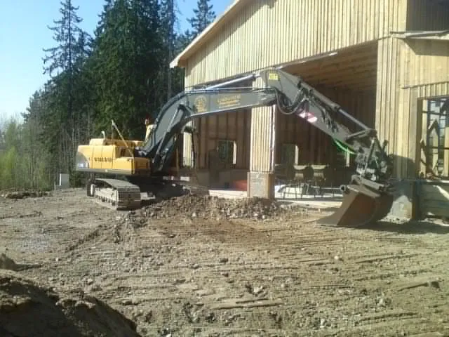 Grävmaskin framför en träbyggnad på en grusplan